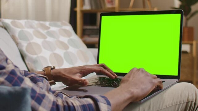 Rear of African American man typing on laptop with green screen while sitting at home. Male in checkered shirt using notebook computer with blank chroma key display on lap indoors. Close up.