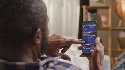 African American man holding smartphone with stock market app open, financial charts and data on screen, sitting at home on couch. Male person tracking investments using mobile trading application. - Powered by Adobe