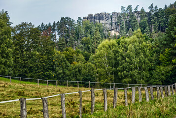 Fototapeta premium Lush green meadow with wooden fence in the foreground and impressive sandstone rock formations in the background, bohemian switzerland national park, czech republic