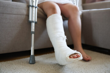 girl with a broken leg in a cast and bandage sits on a sofa metal crutches with height adjustment are at her feet sore leg in focus healthy leg out of focus