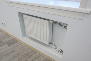 White radiator in a recess under a window, with visible pipes and a grey wood-look floor