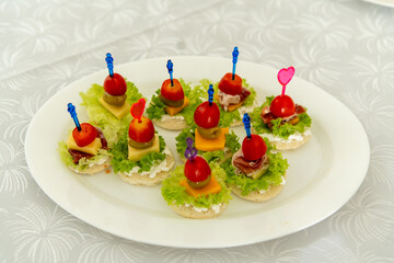 A variety of mini sandwiches topped with cherry tomatoes, cheese, and greens are displayed on a white plate at a cheerful gathering. Decorative picks add a playful touch.