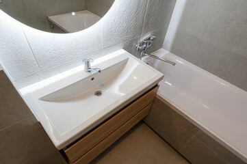 A modern bathroom features a white sink with a wooden cabinet, a round illuminated mirror, a bathtub, and gray tile walls and floors. Chrome fixtures add a sleek touch