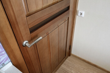 A close-up of a wooden door with a silver handle, next to a white wall with an electrical outlet....