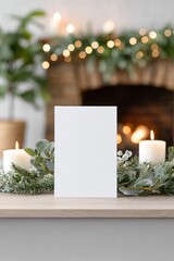 Blank white card mock up with copy space on wooden table, surrounded by greenery and candles, creating a cozy festive atmosphere near a fireplace