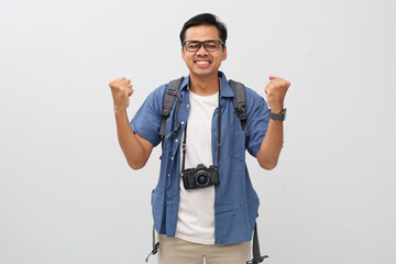 Fototapeta premium A young asian man wearing casual clothes, backpack, camera hanging on neck, making a gesture of celebrating victory by clenching his fist, smiling while closing his eyes, travel or vacation concept.