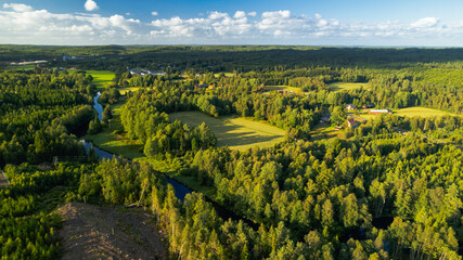 A scenic aerial view of a forested landscape with open fields, winding rivers, and scattered rural houses under a partly cloudy sky. The image captures the vibrant greenery of summer, typical of a Nor