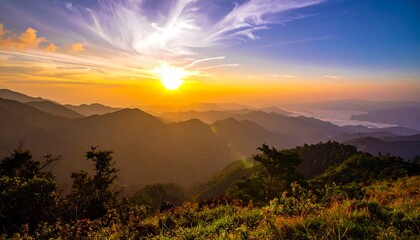 A breathtaking sunrise over a range of lush, rolling mountains. The sky is ablaze with orange, yellow, purple hues