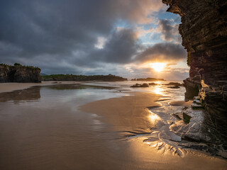 Dramatic cliffs and sandy shore of Playa de las Catedrales in Galicia, Spain, at sunset with golden light breaking through clouds, capturing natural beauty, solitude, and wonder
