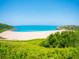 Panoramic view of Playa de Usgo in Cantabria, Spain, featuring turquoise waters, white sand, green cliffs, and a bright blue sky captured on a clear summer day.
