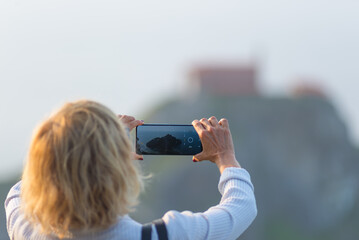 Tourist taking a smartphone photo of San Juan de Gaztelugatxe in Basque Country, Spain, with blurred rocky island and chapel in the background captured on a hazy day