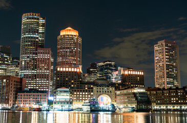 The illuminated downtown Boston skyline, including the Boston Harbor Hotel with its iconic arch, reflecting on the water of the harbor at night. Massachusetts, United States