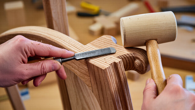 A close-up shot of a person's hands carving wood with a chisel and a mallet. The image shows the intricate and careful work of a woodworker, focusing on the craftsmanship and skill of the artisan.
