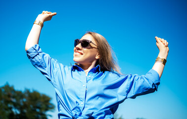 Young woman with blonde hair enjoys the sunny day outdoors, smiling and expressing joy while dressed in a stylish blue shirt against blue sky
