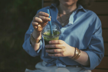 Young woman enjoys a refreshing green smoothie outdoors while dressed in a stylish blue shirt during a sunny day
