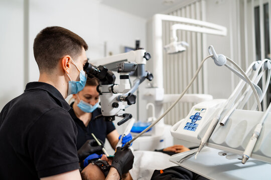 Dental professional uses microscope for precise treatment. A dental professional in a mask examines a patient's teeth with a microscope while a nurse assists during the procedure.