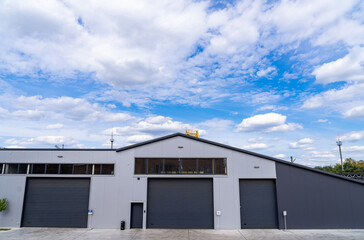Fototapeta premium Modern warehouse under cloudy sky in urban setting. Large warehouse building situated in a city, featuring multiple garage doors and a bright blue sky with clouds.