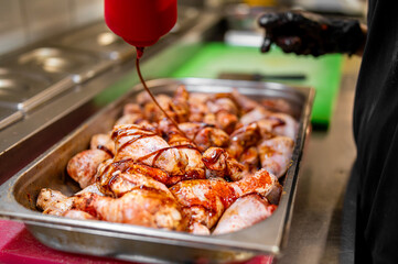 Raw chicken drumsticks being marinated with sauce in a commercial kitchen. Gloved hand applies seasoning. Food prep scene with stainless steel equipment in background