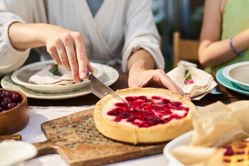 Caucasian young adult man slicing cherry cheesecake with knife while sitting at table outdoors, partial view of woman with bracelet visible, plates and cutlery arranged nearby