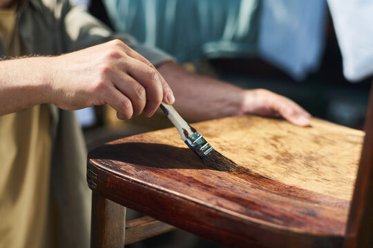 Caucasian middle aged man restoring wooden chair using paintbrush, hand applying varnish to seat surface, focusing on craftsmanship and furniture repair, partial body visible