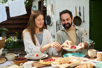 Caucasian young woman and Caucasian middle aged man sitting outdoors sharing dessert at table, smiling and interacting while enjoying food together, surrounded by plates and cups