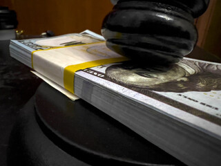 On top of a stack of hundred-dollar bills is a black judge's gavel with a yellow insert on a wooden table. Close-up.