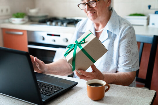 Happy elderly woman holding gift box, using laptop at home kitchen for video call, online connection. Senior virtual celebration alone, holiday greeting, family traditions. Technology lifestyle