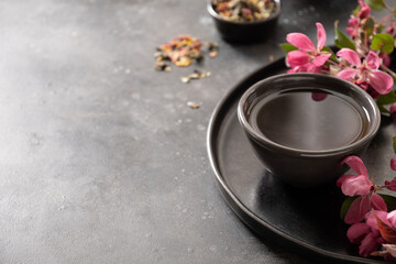Black cast iron teapot and cups with asian tea and pink cherry blossom on black table. Composition with sakura bloom and petals. Japanese style and tea ceremony. Close up. Copy space.