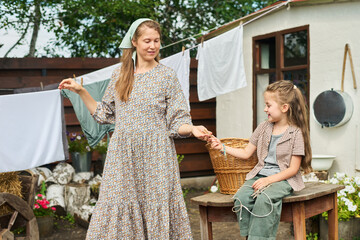 Caucasian middle aged woman hanging laundry outdoors while smiling at Caucasian child sitting on wooden table holding wicker basket, both interacting in backyard setting