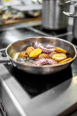 Close-up of octopus tentacles and potato slices frying in oil on a stainless steel pan over an electric stove in a professional kitchen setting.
