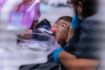 A dentist in purple scrubs examines a young boys teeth while he sits in the dental chair, wearing protective glasses and gloves during the procedure.