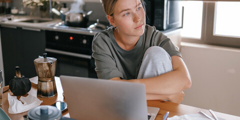  Gen Z woman working from home at cozy kitchen table with laptop and phone. Digital nomad...