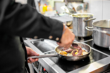 Professional chef cooking octopus and vegetables in a frying pan on stovetop in commercial kitchen. Culinary preparation, seafood dish, gourmet cuisine.