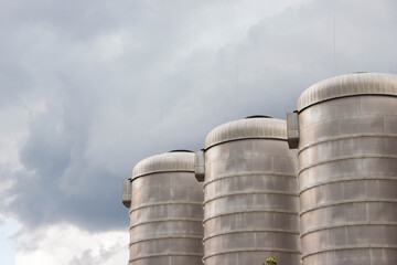 Three large metallic silos stand against a dramatic cloudy sky, showcasing industrial architecture and storage capabilities in a rural landscape with a hint of nature
