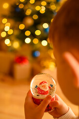 Vertical photo of children’s hands gently holding a Christmas snow globe against glowing...