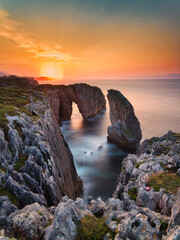 Coastal archway at sunset on a rugged shoreline - A breathtaking sunset at a rocky beach with a natural arch and a sea stack, with soft water and a vibrant orange sky.