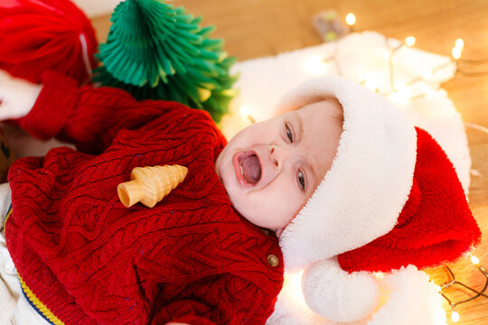 Adorable baby boy in santa hat and with wooden toy crying while lying under festive christmas tree and lights. Sad infant overwhelmed, too emotional holidays.