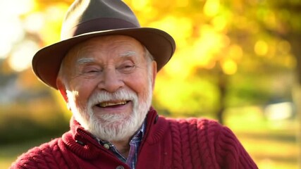 Smiling elderly man with white beard wearing fedora and red sweater, blurred autumn foliage in the background - Powered by Adobe