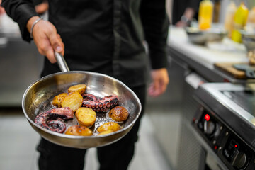 Chef holding pan with grilled octopus and roasted potatoes in professional kitchen. Gourmet seafood dish, freshly cooked and served in stainless steel cookware.