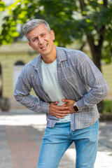 Obraz premium Caucasian man traveler feeling sudden strong abdominal stomach ache, gastritis problem. Upset tourist having symptom poisoning diarrhea indigestion peptic ulcer pancreatitis on city street. Vertical.
