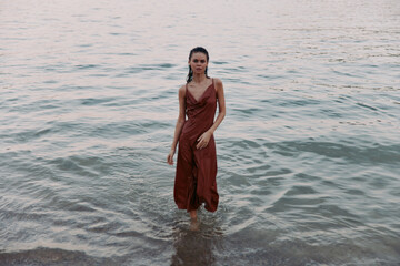 A woman stands in shallow sea water wearing a rust dress, posed with calm expression as gentle waves lap the shore, creating a minimalist fashion portrait.
