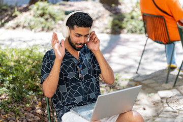 Fototapeta premium Indian bearded young man using laptop having video call chat conversation for online meeting and presentation outdoors. Happy male tourist in headphones recording blog sitting in urban city street.