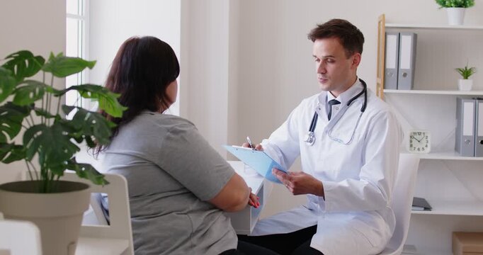 Male physician or cardiologist giving consultation to overweight fat female patient in clinic about wellness, nutrition, weight loss, answering questions, giving advices and describing treatment plan.