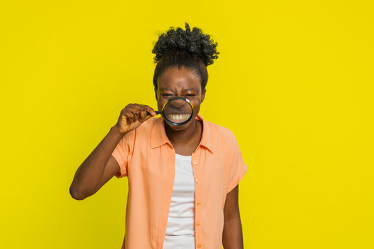 African American woman holds magnifying glass distorting her smile, creating comical effect. Black playful girl on yellow background making funny silly facial expressions grimacing face fooling around