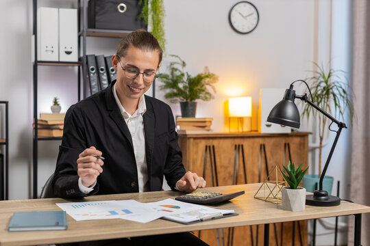 Focused young office worker working with schedules and financial graphics while using calculator sitting at home office desk. Happy stylish businessman freelancer compare and analyze graphs charts.