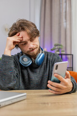 Sad displeased man use smartphone typing browsing, loses becoming surprised sudden lottery game results, bad news, fortune loss fail, deadline, virus. Young adult guy at home office at table. Vertical