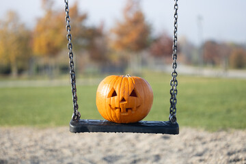 Jack-o'-lantern pumpkin with carved face sits on a swing in a playground, surrounded by autumn...