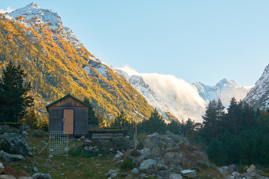 A charming rustic cabin nestles in a high-altitude autumn valley, its wooden walls standing in warm contrast to the snow-capped peaks towering behind.