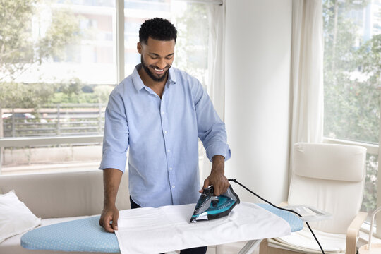 Smiling gen z male homeowner ironing clothes in living room - Powered by Adobe