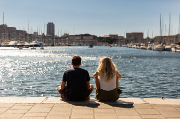 Jeune couple assis sur le quai du port de Marseille regardant l’eau, vue de dos face au soleil, bateaux, ville et ciel bleu en arrière-plan, Vieux-Port de Marseille